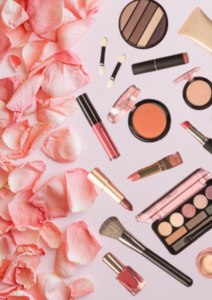 Pink makeup products including lipstick, eyeshadow, blush, brushes, and nail polish arranged with rose petals