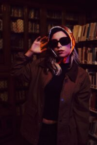 Woman wearing sunglasses, a colorful headscarf, brown jacket, and black outfit in a vintage library