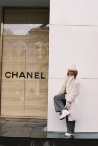 Woman in a white winter coat, beanie, scarf, and boots posing outside a Chanel store